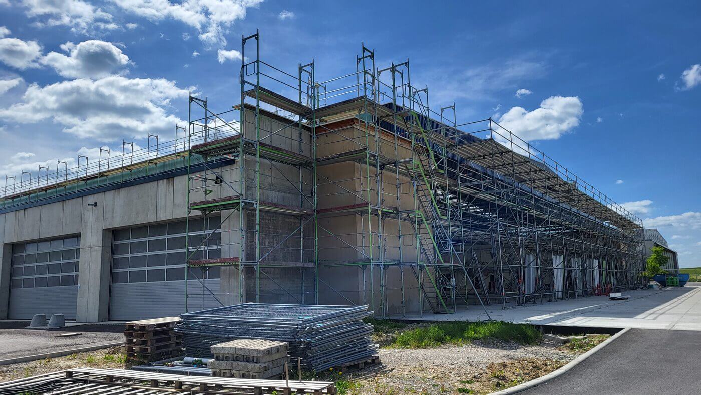 Große Halle mit eingerüsteter Fassade, mehreren geschlossenen Rolltoren und Materialstapeln.